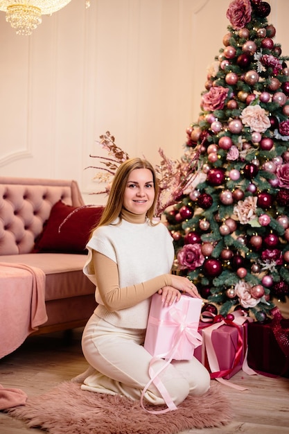 Charming woman dressed in a white wool suit holds a pink gift against the background of a decorated Christmas tree in a room next to a New Year tree and a pink sofa gifts and candles