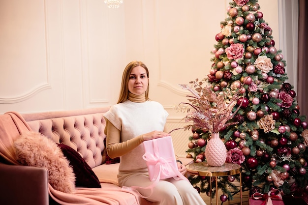 Charming woman dressed in a white wool suit holds a pink gift against the background of a decorated Christmas tree in a room next to a New Year tree and a pink sofa gifts and candles