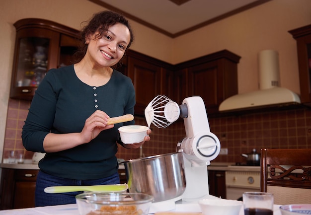 Charming woman housewife pastry chef holding mascarpone cheese and biscuit cookies smiles looking at camera while preparing homemade tiramisu dessert in home kitchen