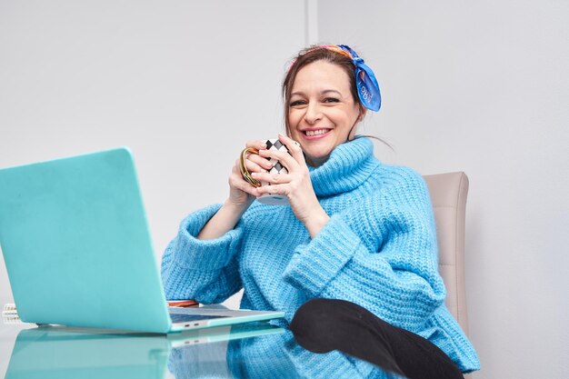 Charming woman sitting in the living room using her laptop while having a cup of coffee looking at the camera