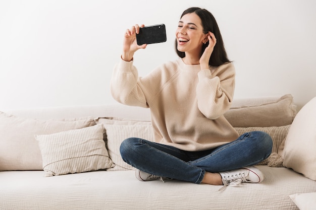 charming woman smiling and holding cell phone while sitting on sofa at home