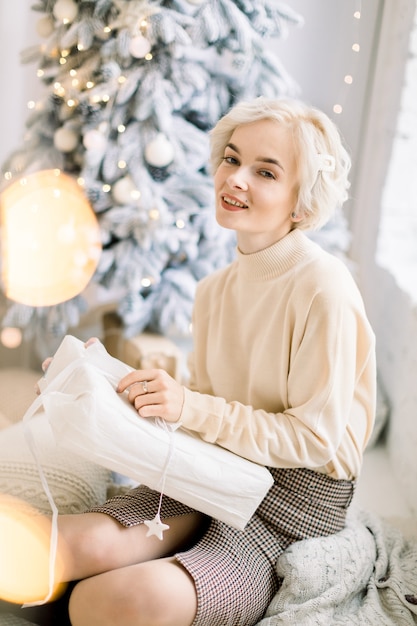 Charming woman in sweater and skirt posing in light decorated room with Christmas gift