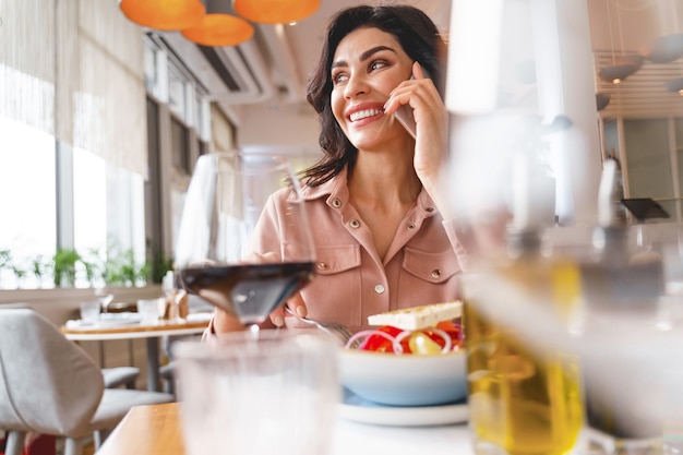 Charming woman talking on cellphone and smiling while sitting at the table with food and wine