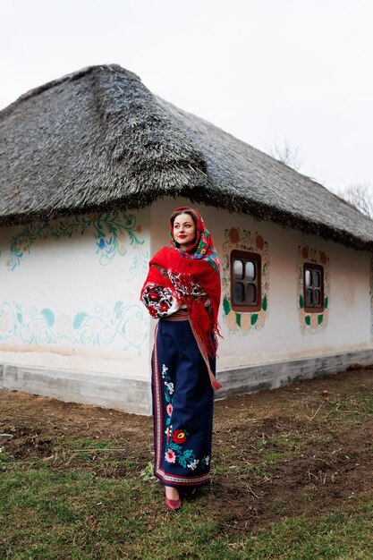 Charming woman in traditional ukrainian handkerchief necklace and embroidered dress standing at background of decorated hut Ukraine style folk ethnic culture