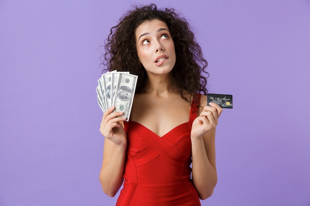 お金とクレジットカードのファンを保持し、紫色の壁に孤立して立っている赤いドレスを着ている魅力的な女性