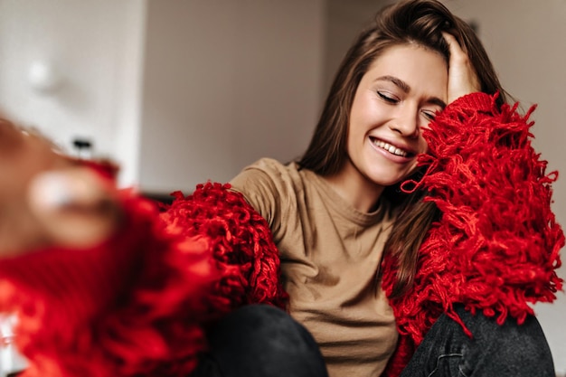 Charming woman with gloss on her lips makes selfie Snapshot of girl with closed eyes dressed in knitted red sweater and beige top