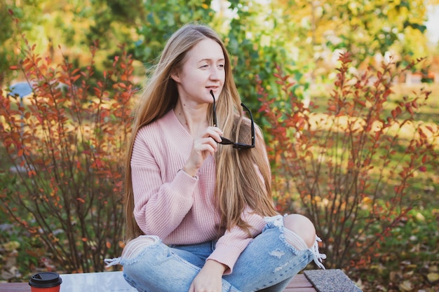 Charming woman with long hair and in casual sunglasses sits on bench in beautiful autumn park