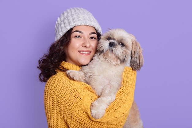 Charming woman with Pekingese puppy, cuddles gently near face, wears knitted sweater and cap.