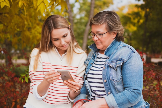 Charming women mom and daughter are sitting on bench in beautiful autumn park and looking at photos on smartphone