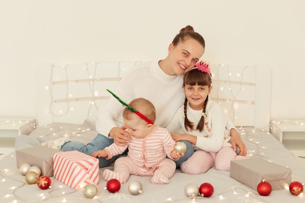 Charming young adult woman lovely mother with her children wearing casual attires, sitting on bed and hugging each other, enjoying winter holidays, expressing happiness.