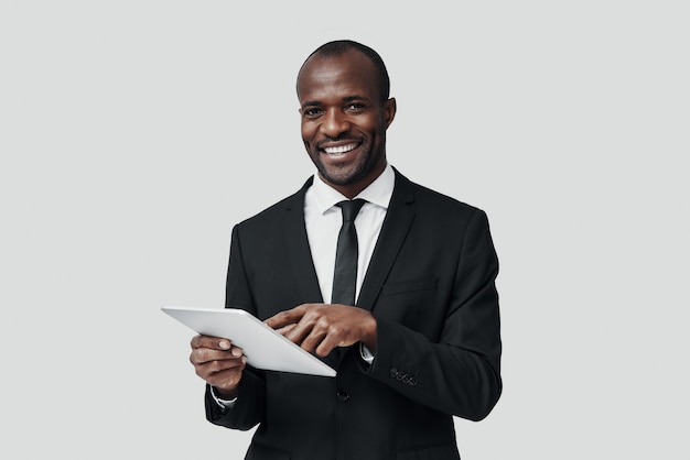 Charming young African man in formalwear working using digital tablet while standing against grey wall