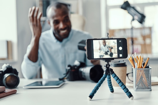 Photo charming young african man in shirt showing digital camera and telling something while making social media video
