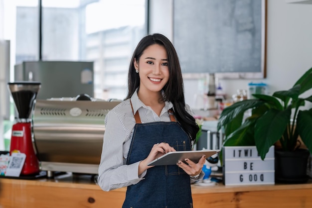 コーヒーショップのカウンターの前でタブレットを保持しているエプロンを身に着けている魅力的な若いアジアの実業家の所有者。カメラを見てください。