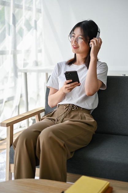 A charming young Asian woman enjoys the music on her headphones while sitting on a sofa