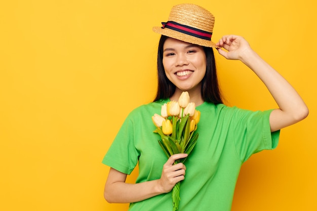 魅力的な若いアジアの女性の緑のTシャツは、帽子の孤立した背景に黄色の花の花束を変更せずに