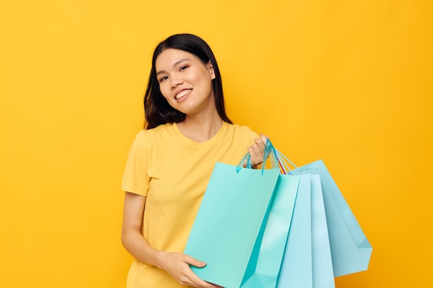 Charming young Asian woman in a yellow Tshirt with multicolored shopping bags yellow background unaltered