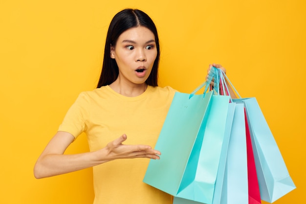 Charming young Asian woman in a yellow Tshirt with multicolored shopping bags yellow background unaltered