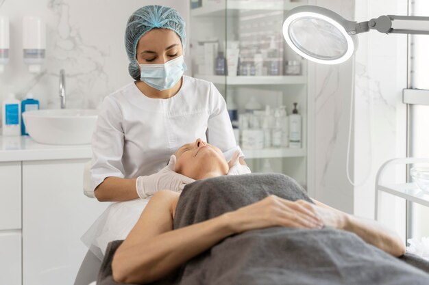 Charming young beautician in white lab coat examining her senior female patient face