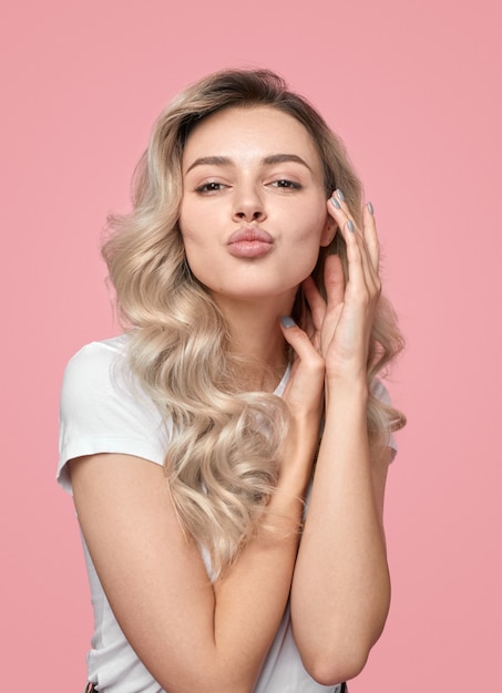 Charming young blonde female model with long wavy hair and pout lips sending kiss while looking at camera against pink background