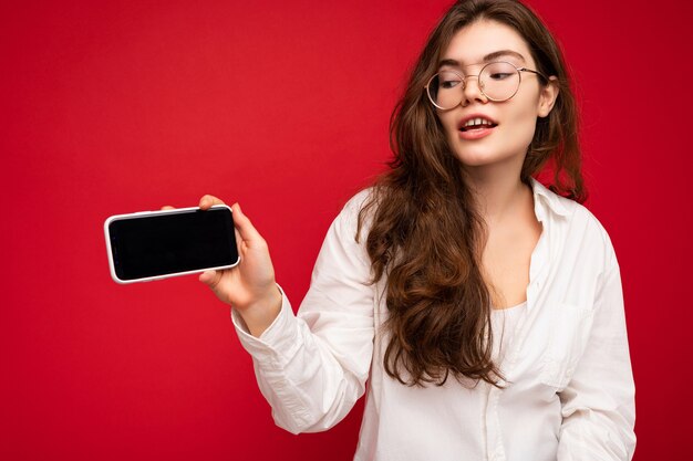 Charming young brunet woman wearing white shirt and optical glasses isolated over red