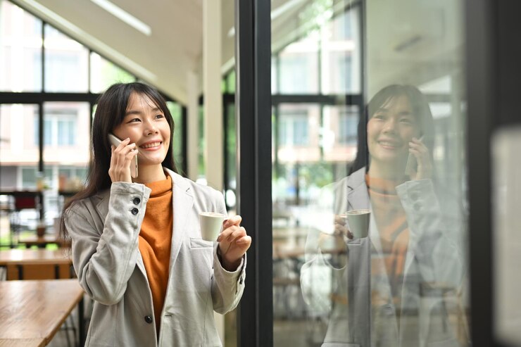 オフィスの窓の近くに立って愉快な電話会話をしている魅力的な若いビジネスウーマン