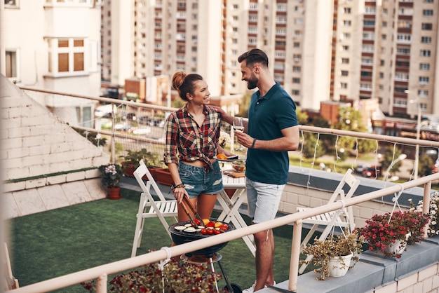 Очаровательная молодая пара в повседневной одежде готовит барбекю и улыбается, стоя на патио на крыше на открытом воздухе