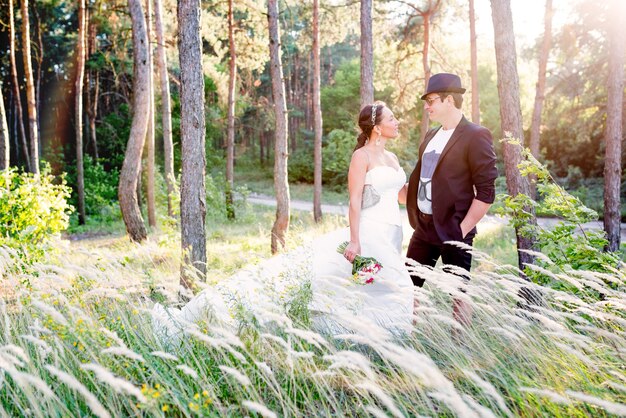 Charming young couple just married beautiful woman in a white dress and groom in a suit and hat posing in a field with tall grass.