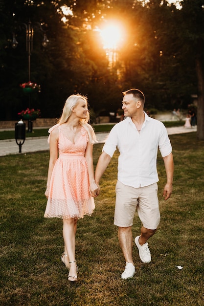 Charming young couple walking and looking to each other. Romantic couple dating holding hands against sunset outdoor.