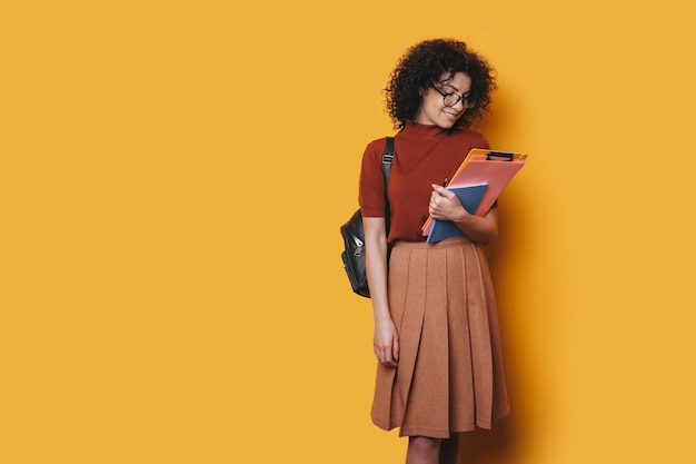 Charming young curly female student looking at her studying books isolated on yellow background.