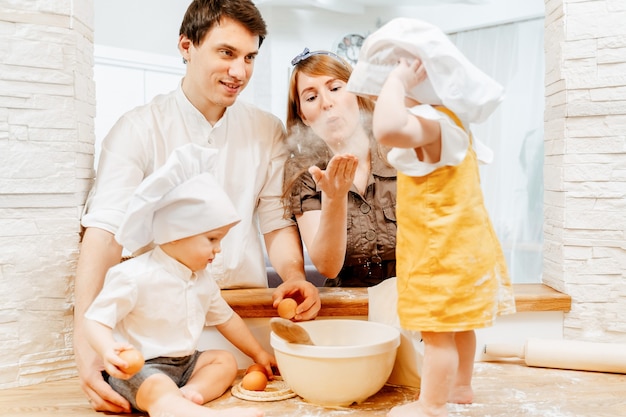 Charming young family dad mom and two year old brother and sister in cook clothes are preparing dough for a pie in a cozy kitchen. Weekend family home together concept