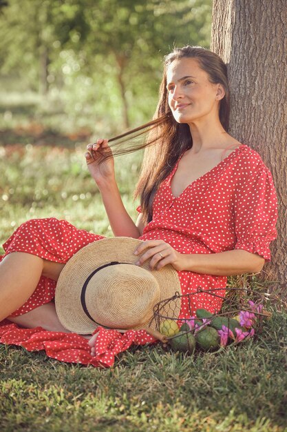 Photo charming young female in red dress and straw hat sitting on grass