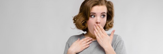 Charming young girl in a gray T-shirt on a gray background. The girl covered her mouth with her hand