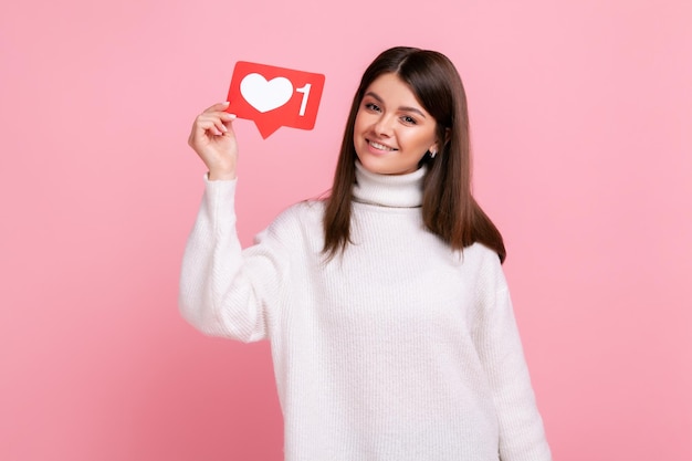 Charming young girl holding heart like icon of social media and looking at camera with toothy smile