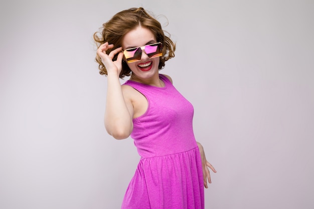 Charming young girl in pink dress on gray background. Cheerful girl in square glasses. The girl adjusts glasses