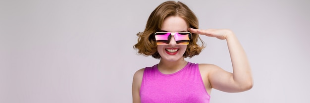 Charming young girl in pink dress on gray background. Cheerful girl in square glasses. The girl put her hand to her head