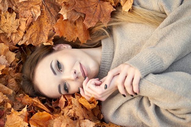 Charming young girl with cute smile liying in the autumn leaves and enjoying it, holding hands near face and smiling. Top view.