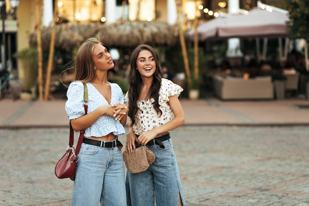 Charming young girls in florals colorful blouses and stylish denim pants walk in city and smile sincerely outdoors