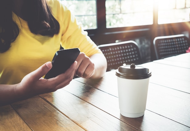 魅力的な若いhipsterの女の子の手は、コーヒーショップの木製テーブルに座って彼女のスマートフォンで使用しています。アジアの女性はカフェに座ってリラックス - ヴィンテージカラースタイル。