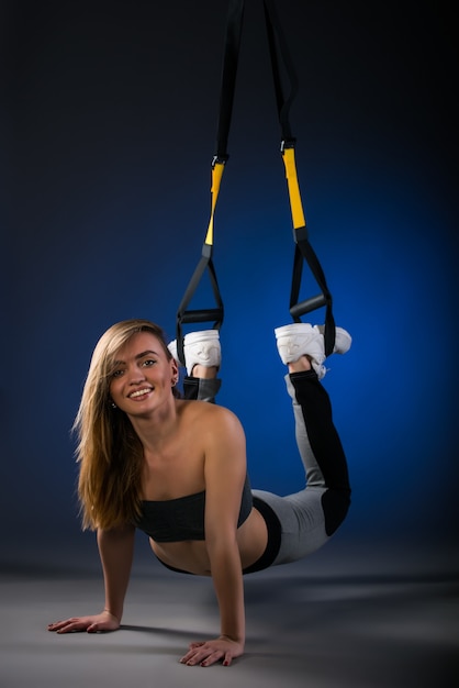 Affascinante giovane modello di fitness caucasico positivo dai capelli lunghi che fa flessioni dal pavimento con l'aiuto di cinghie sospese sul blu