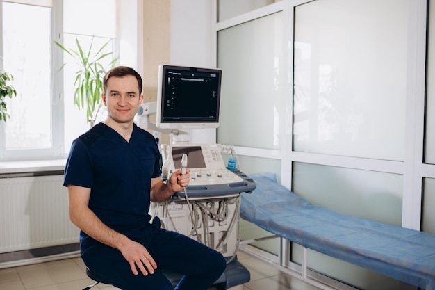 Charming young male doctor smiling to the camera sitting at his office near ultrasound scanning machine Handsome friendly doctor enjoying working at the hospital copy space Ultrasonic concept