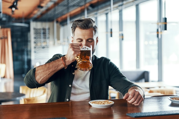 Charming young man in casual clothing drinking beer while spending time in the pub