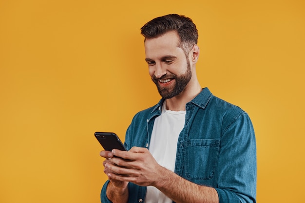 Charming young man in casual clothing using smart phone and smiling while standing  