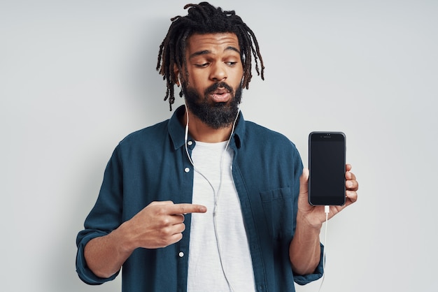 Charming young man in casual wear pointing copy space and listening music while standing against grey wall