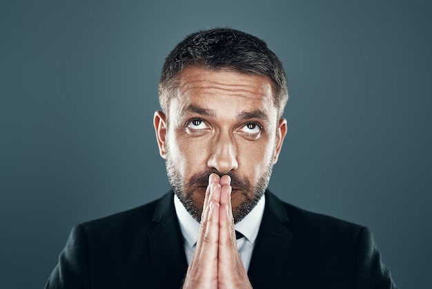 Charming young man in full suit keeping hands clasped while standing against grey background