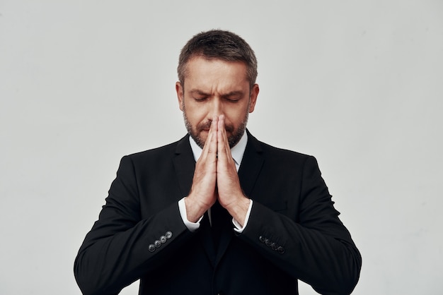 Charming young man in full suit keeping hands clasped while standing against grey background