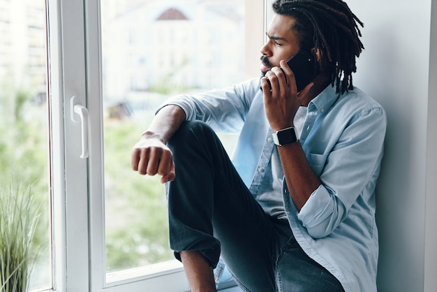 Charming young man talking on the smart phone while sitting on the window sill indoors