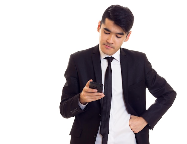 Charming young man with black hair in white shirt and black suit with tie using his phone