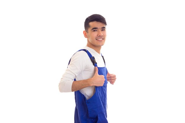 Charming young man with dark hair wearing in white shirt and blue overall showing thumb up