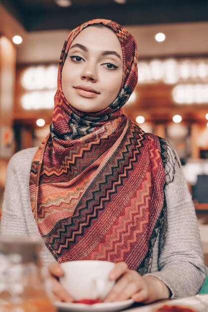 Charming young Muslim girl in hijab drinking fragrant coffee and looking at camera