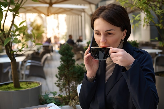 Photo charming young successful brunette woman drinking coffee and talking on a cell phone while relaxing outdoors during a coffee break on a beautiful sunny day